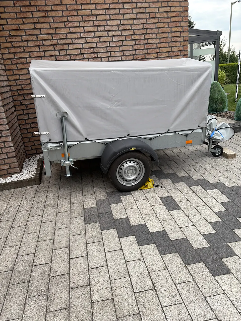 Humbaur-Anhänger mit grauer Hochplane auf gepflasterter Fläche; Seitenwand, Rad und Deichsel sind sichtbar.