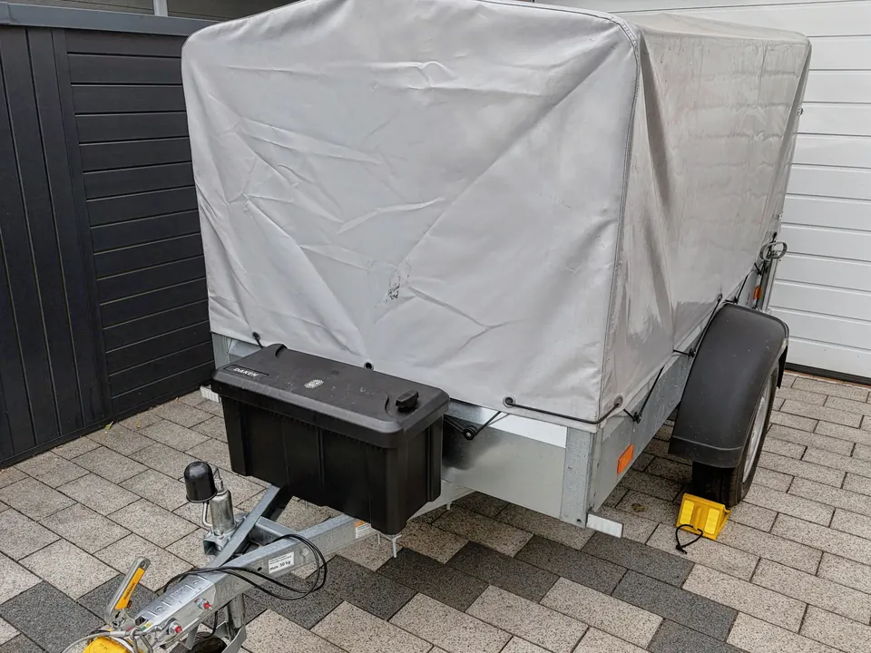 Der Humbaur-Anhänger schräg von vorn mit grauer Hochplane, Deichsel und aufgesetztem schwarzen Kasten.